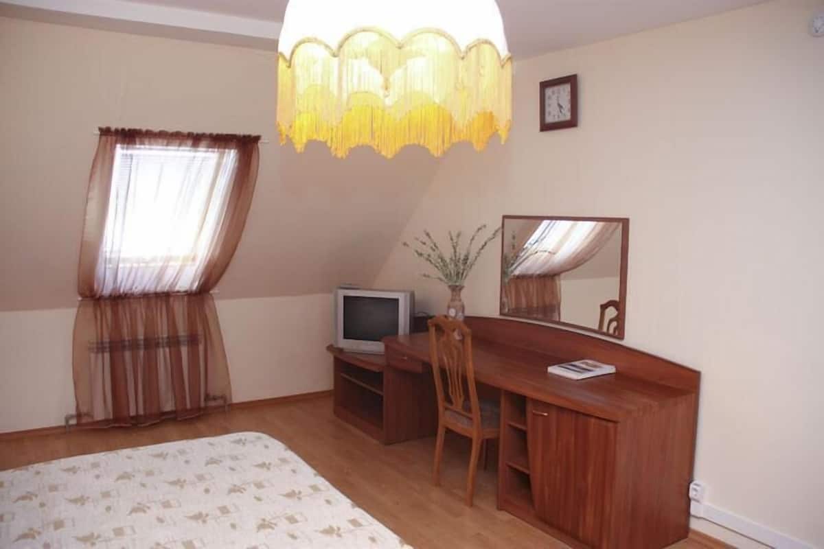 A bedroom with a bed, a desk, a chair, a television, a mirror, and a window with curtains.