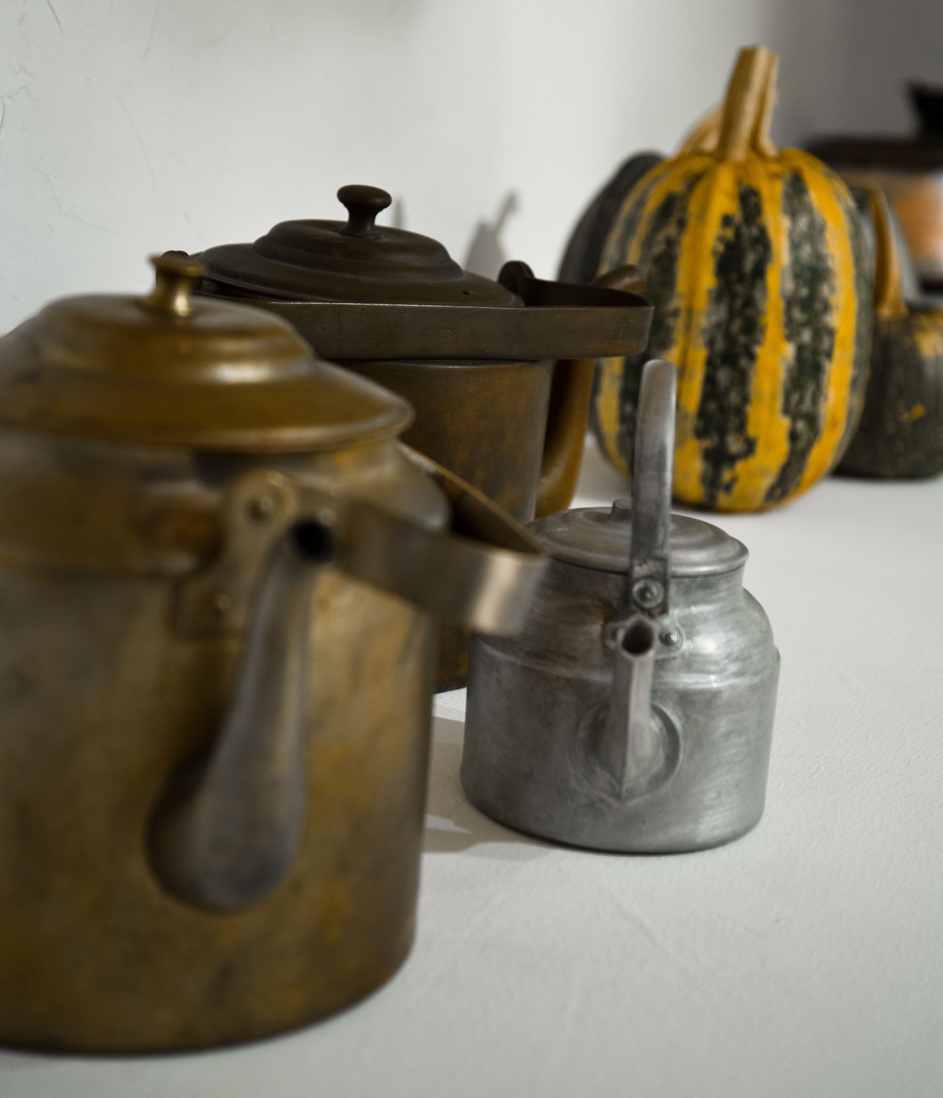 A close-up of a brass teapot with a handle and a spout, a silver teapot with a lid and a handle, and a pumpkin with stripes in the background.