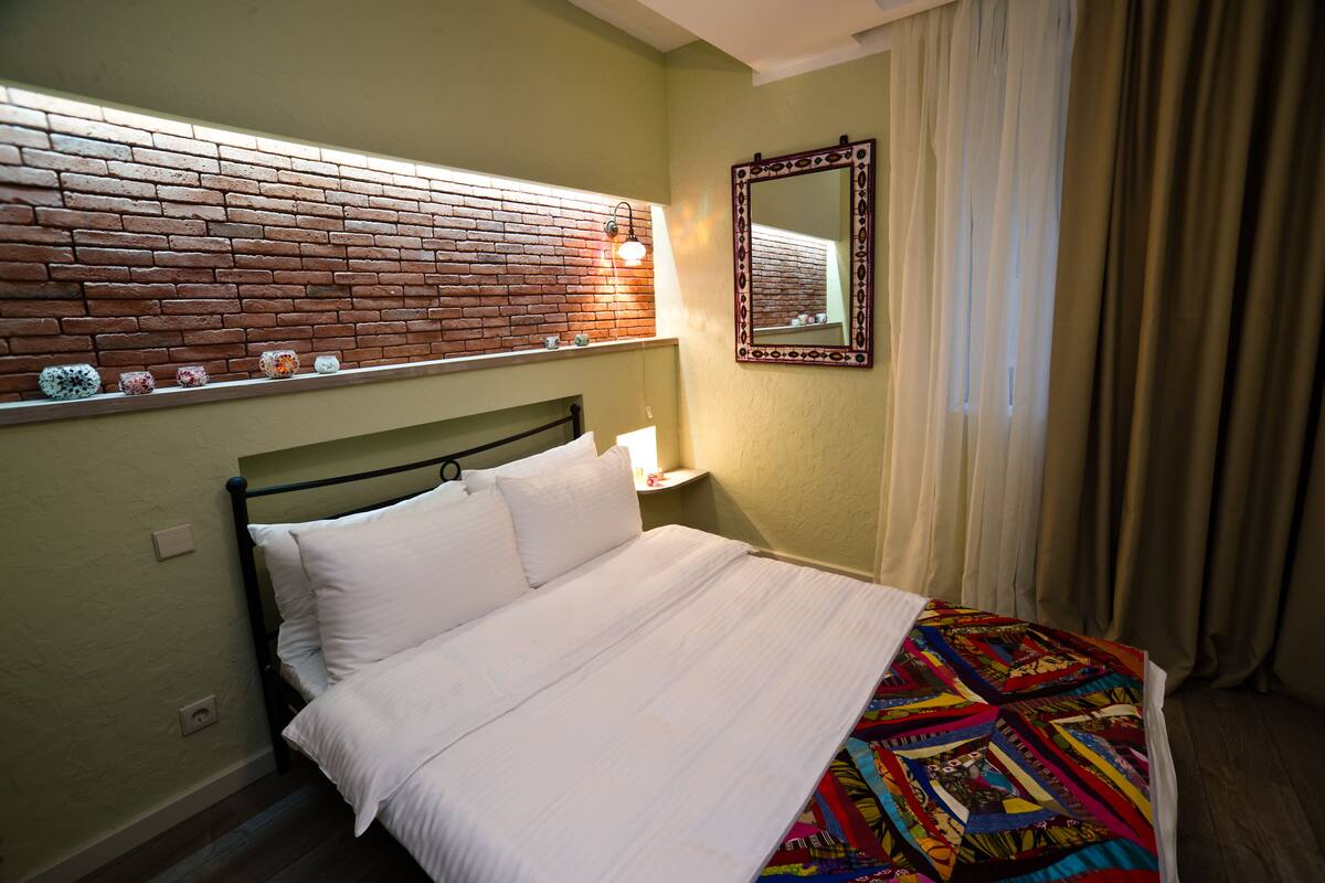Un lit bien fait, avec du linge de lit blanc, une couverture colorée, un mur décoré d’un motif en briques, un miroir et une table de chevet avec une lampe.