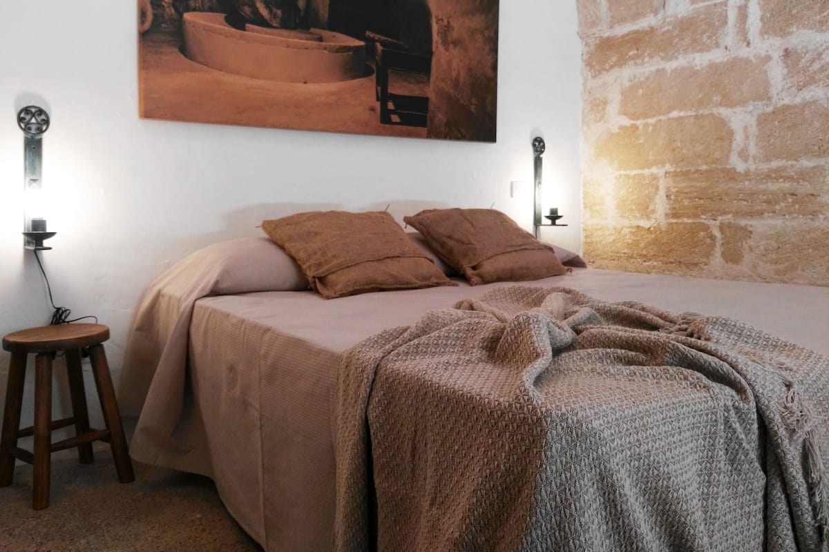 Une chambre à coucher avec un lit, deux oreillers, une couverture, un petit tabouret et un tableau accroché au mur.