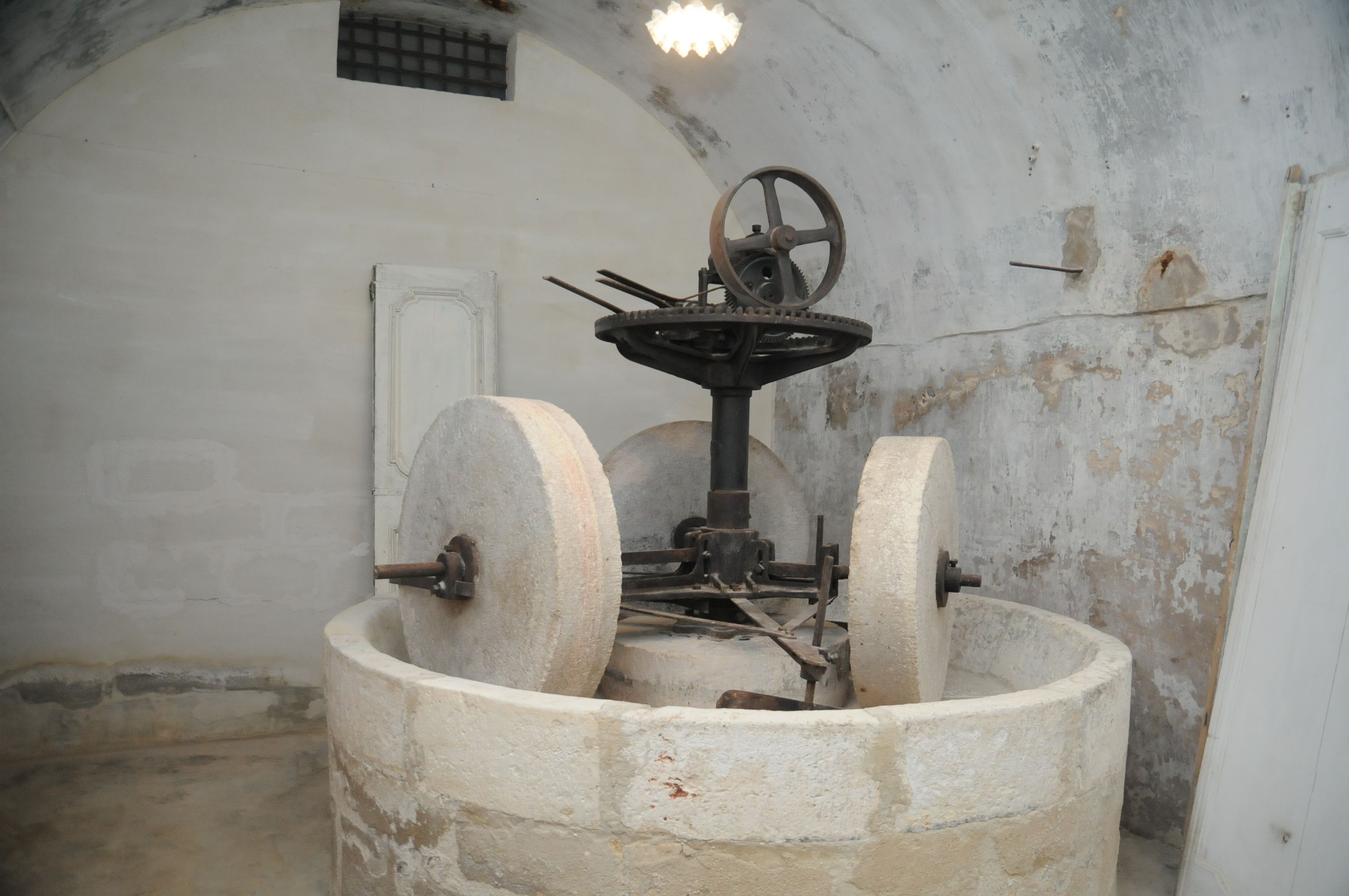 A large, circular stone structure with a central wheel mechanism, possibly a historical water pump, in a room with white walls and a small window.