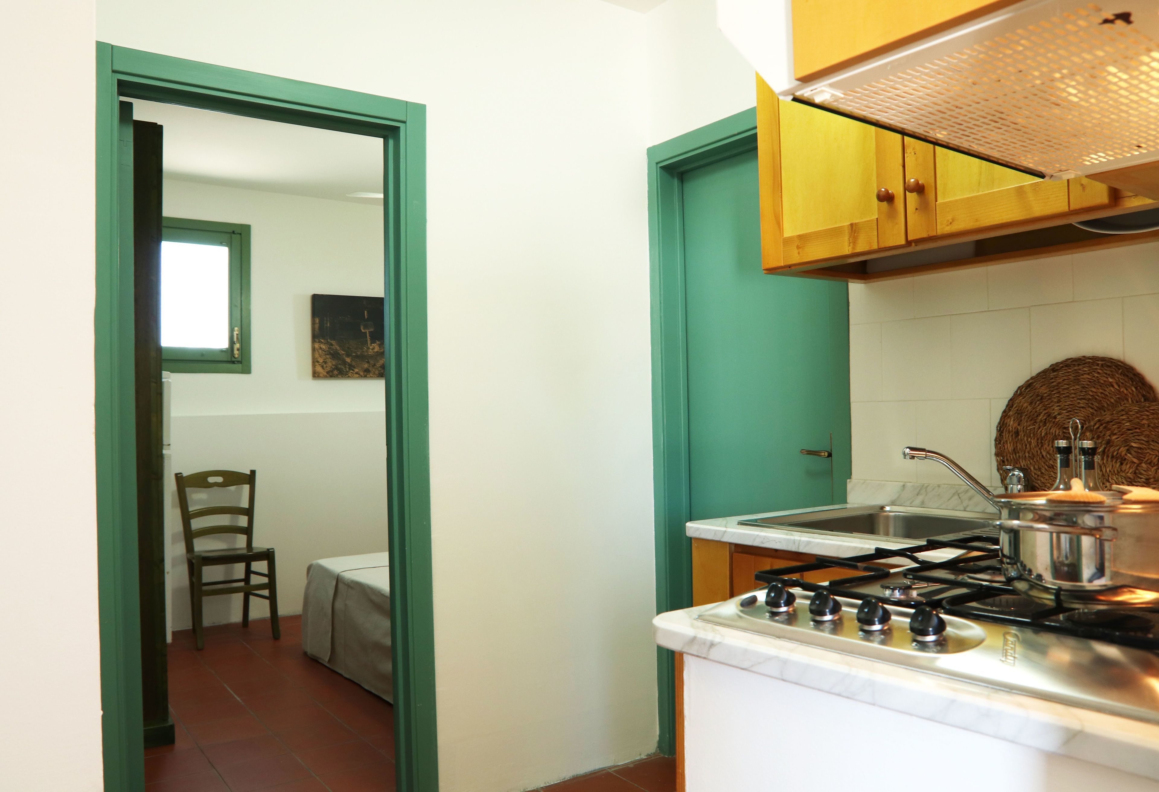A kitchen with a stove, sink, and wooden cabinets. There is a chair and a table in the adjacent room.