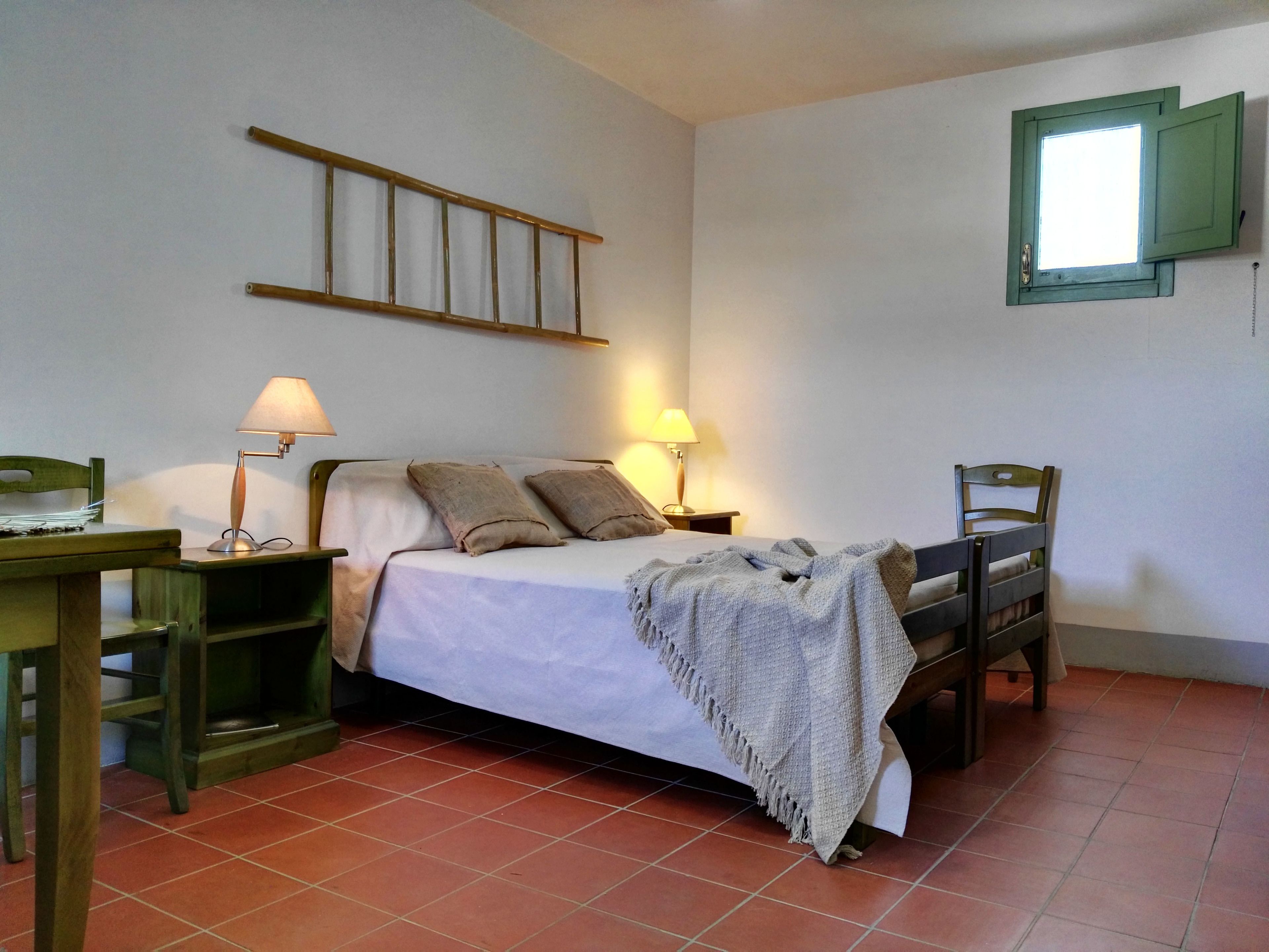 Een slaapkamer met een bed, twee bedlampen, een houten hoofdbord, een stoel, een klein tafeltje en een raam met een groene luik.