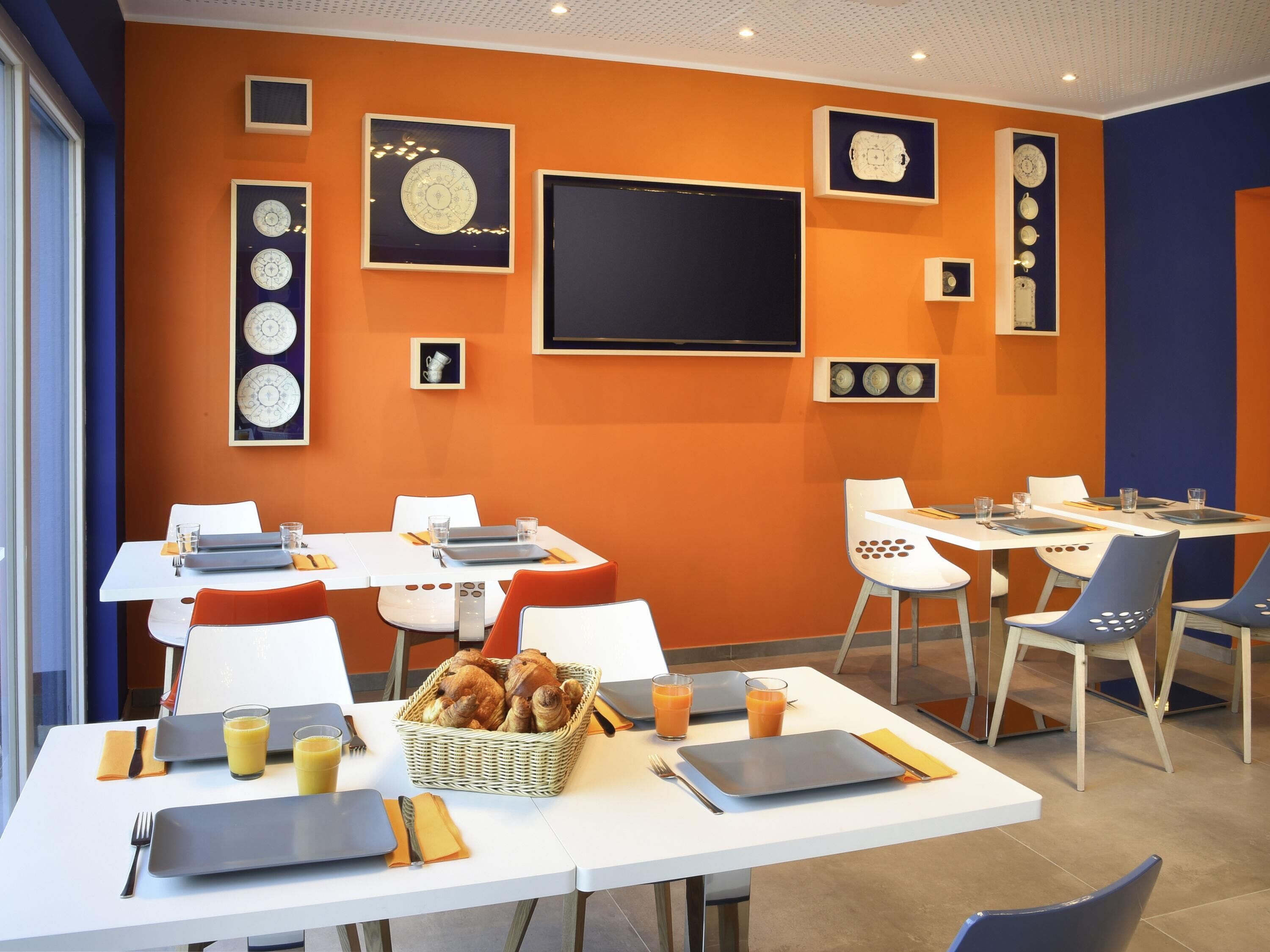 A dining area with white tables, orange walls, and a blackboard. There are plates, glasses, and a basket of bread.