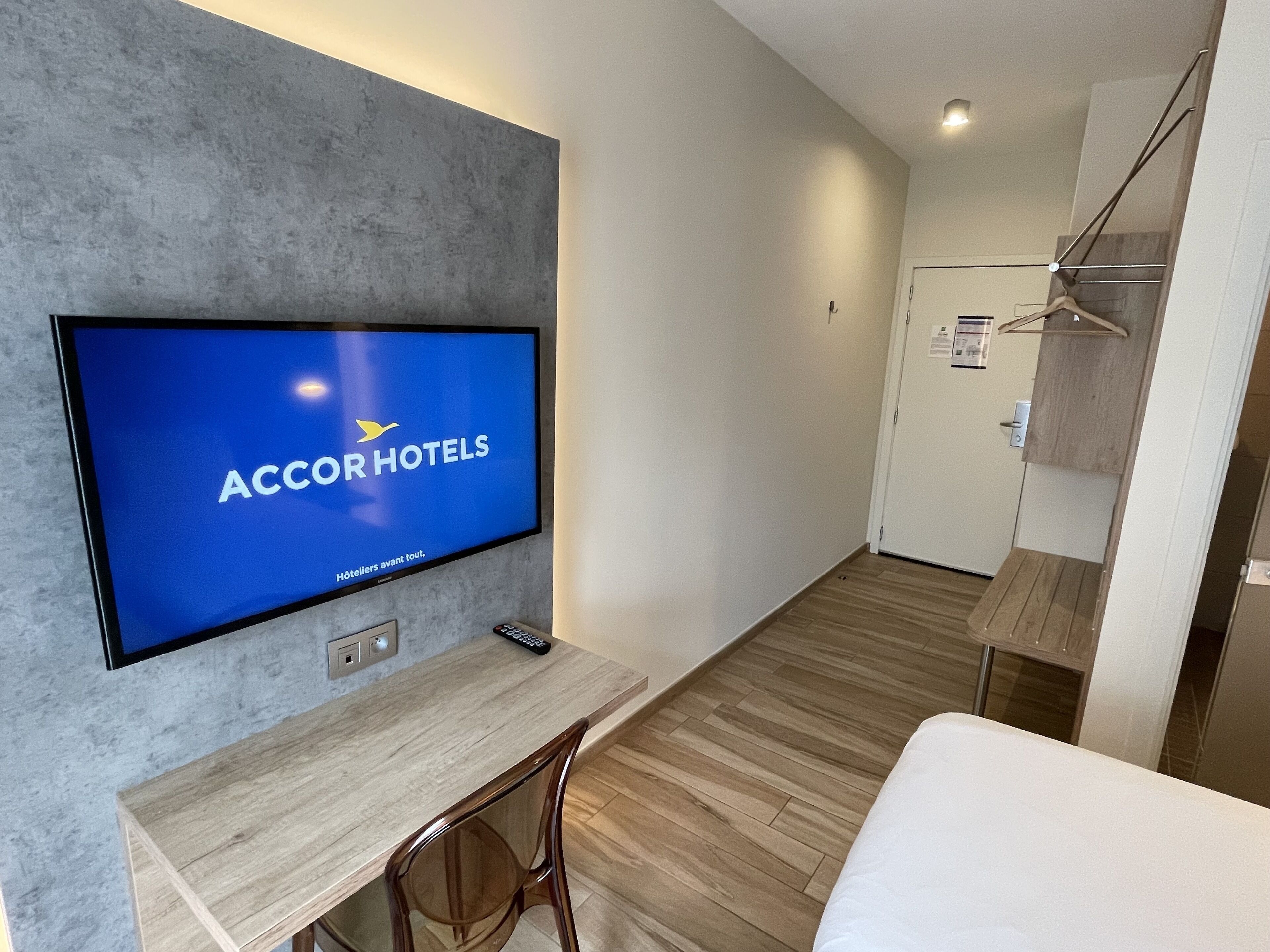 A modern hotel room with a flat-screen TV displaying ACCORHOTELS, a wooden desk, a chair, and a bed.