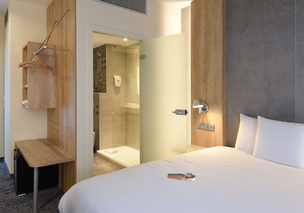 A hotel room with a bed, a wooden side table, a hanging coat rack, a shower area, and a door with a handle.