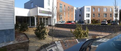 Un edificio moderno con techo plano y ventanas amplias, rodeado de coches aparcados y un pequeño jardín.