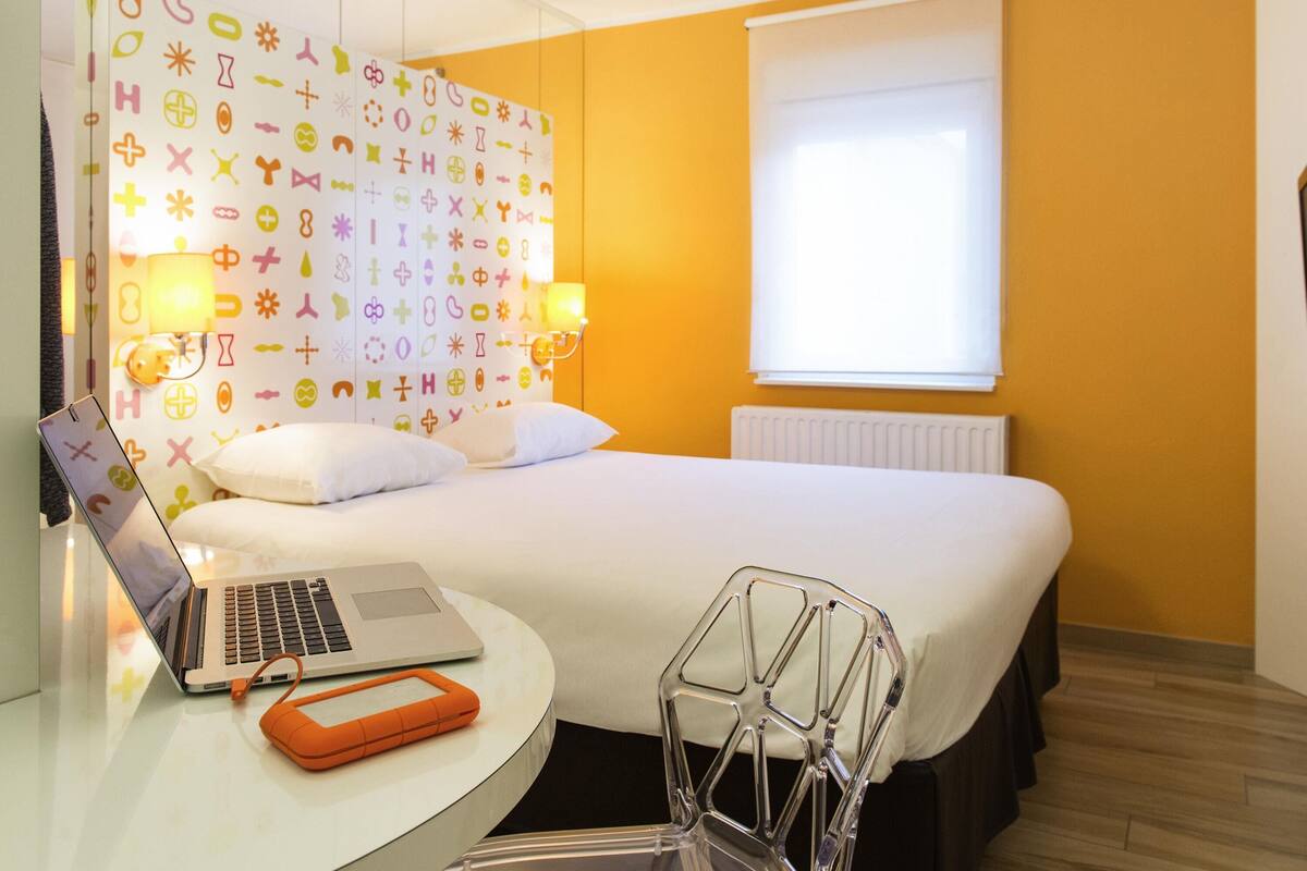 A hotel room with a bed, a desk with a laptop, a chair, and a window with blinds.