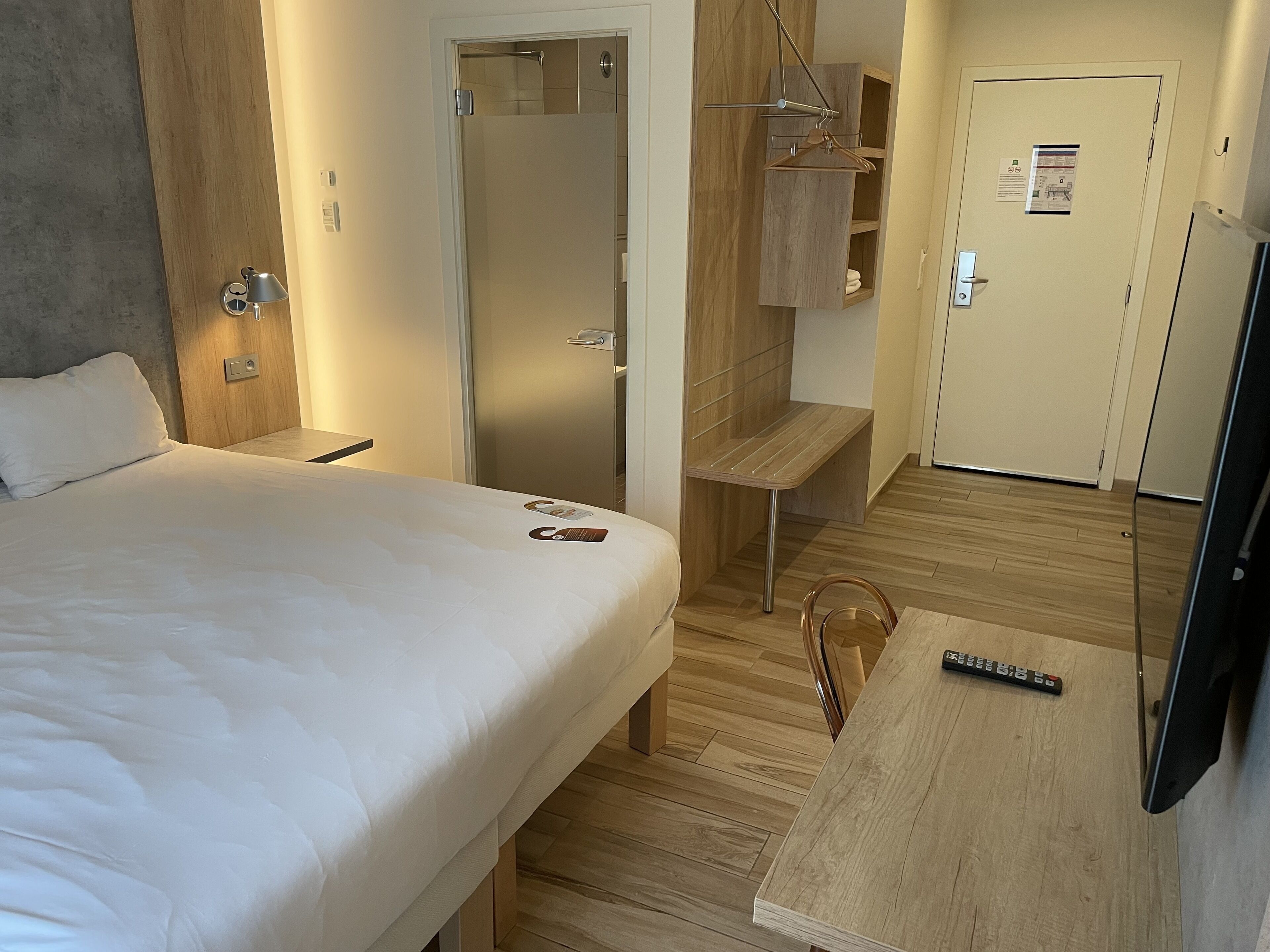 A hotel room with a bed, a wooden bench, a mirror, a wooden shelf, and a door with a towel rack.
