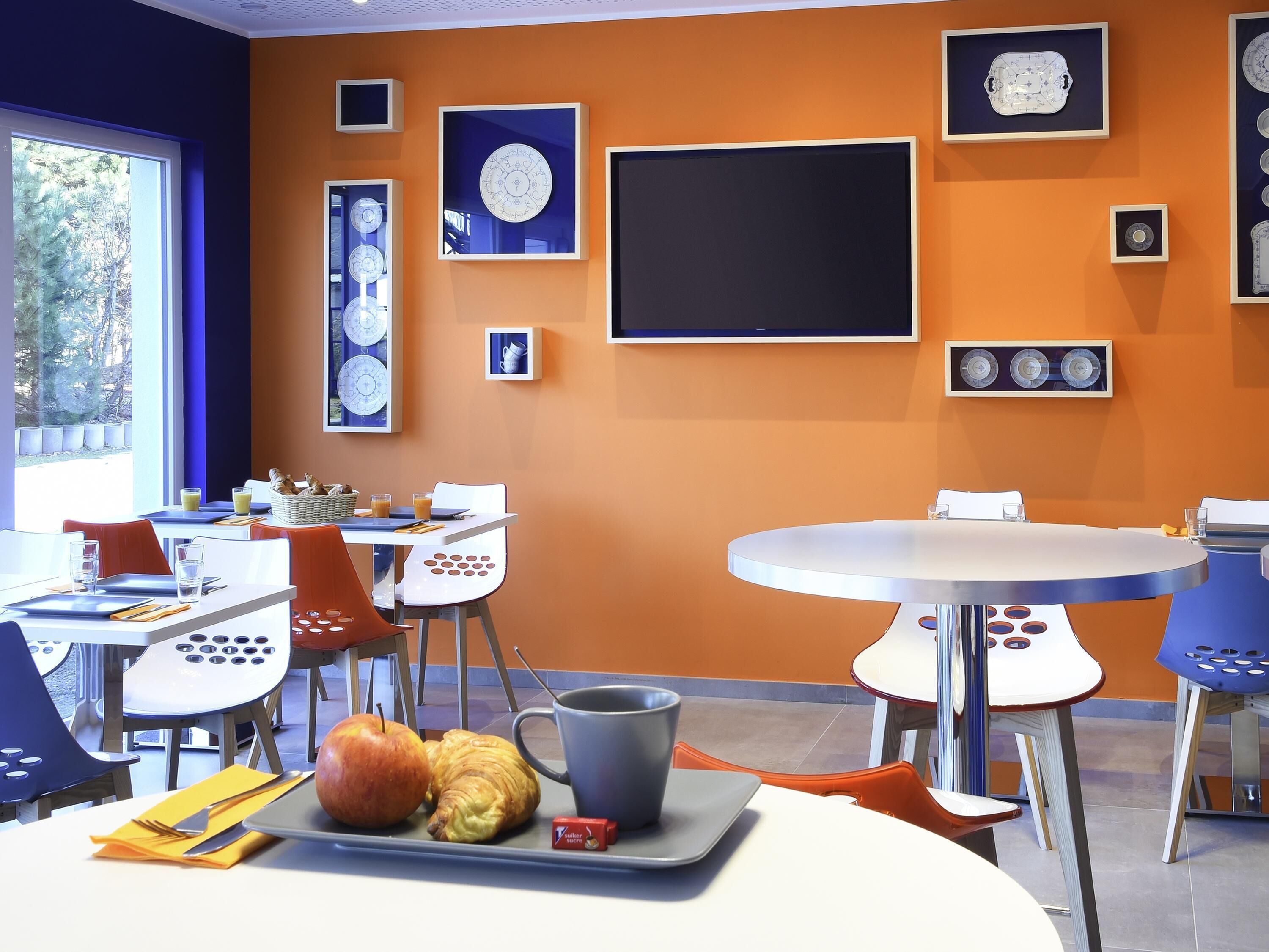 Ein moderner Essbereich mit orangefarbenen Wänden, weißen und blauen Stühlen sowie unterschiedlichen Tischdeckungen, darunter Teller, Gläser und ein Croissant.