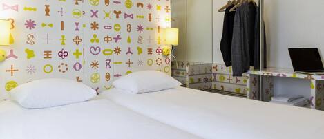A hotel room with a bed, two pillows, a bedside table with a laptop, a mirror, and a wall with colorful patterns.