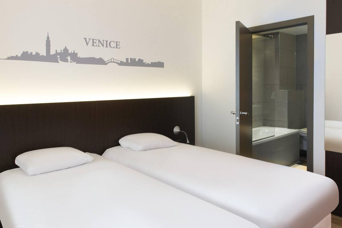 A hotel room with two beds, a city skyline mural, a shower, and a door to the bathroom.