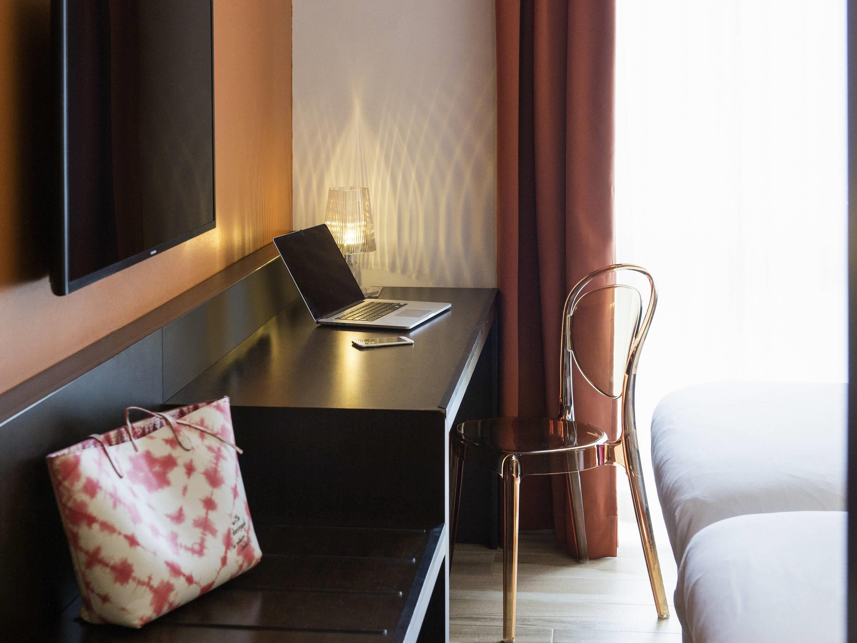 A hotel room with a desk, a laptop, a smartphone, a chair, a handbag, and a bed.
