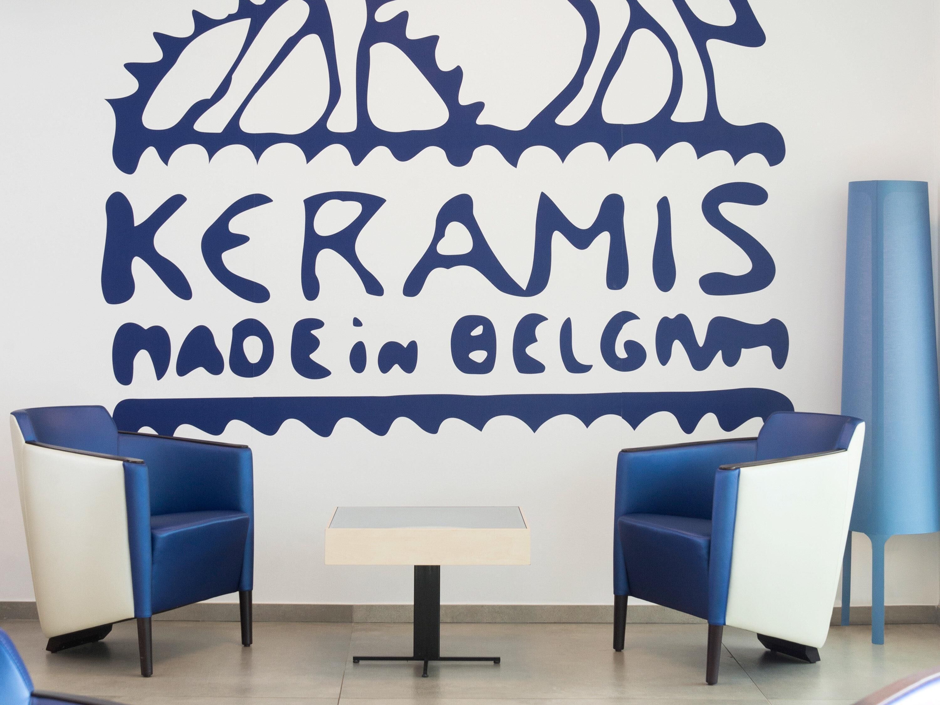 Un intérieur moderne avec un mur orné de l’inscription « KERAMIS, FABRIQUÉ EN BELGIQUE » et deux chaises bleues disposées autour d’une petite table placée au centre.
