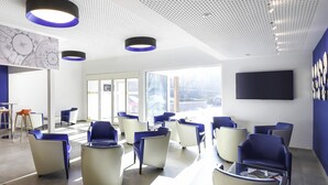 Une salle d’attente moderne, dotée de chaises bleues et blanches, d’un téléviseur à écran plat et de grandes fenêtres offrant une vue sur la ville.