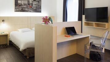 A hotel room with a bed, a desk with a laptop, a TV, and a clear chair.