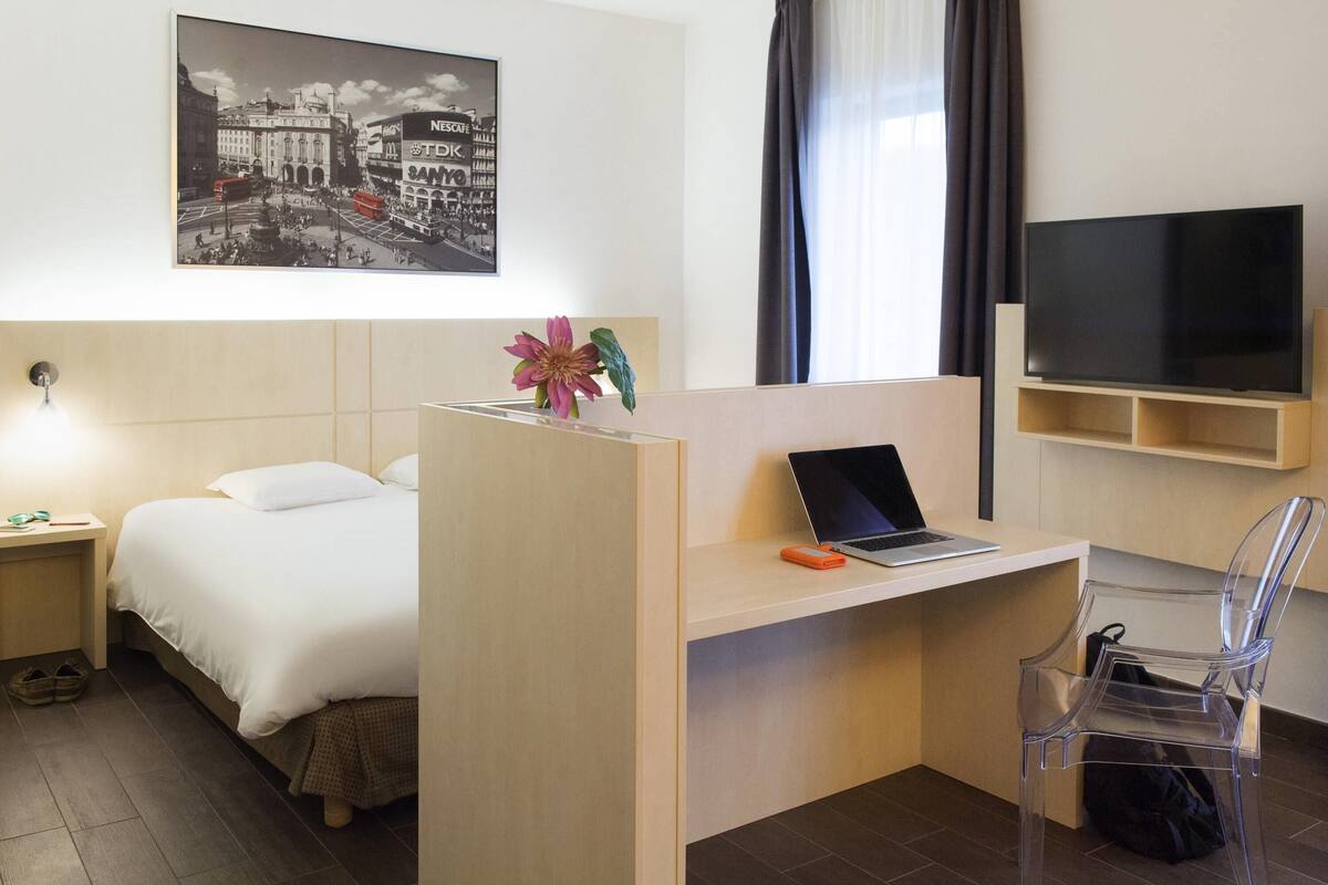 A hotel room with a bed, a desk with a laptop, a TV, and a picture of a cityscape.