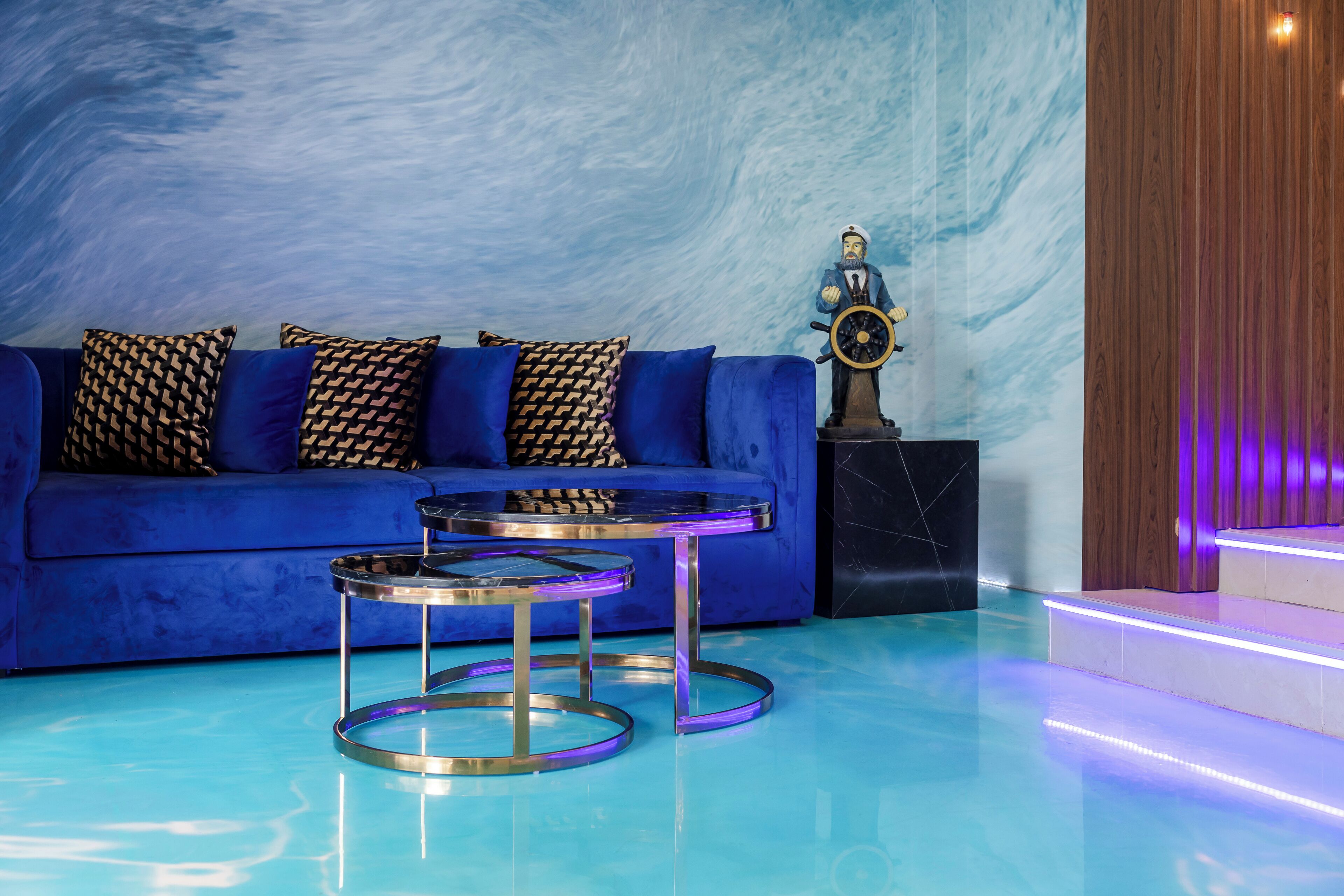 Ein modernes Wohnzimmer mit einem blauen Sofa, gemusterten Kissen, zwei Tischen mit Glasplatte und einer Statue eines Schiffsschiffsrades.