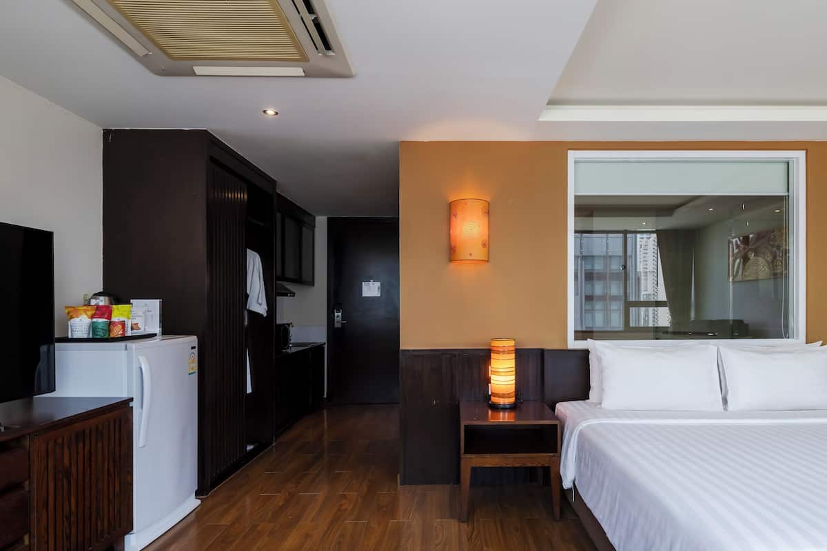 Ein Hotelzimmer mit Bett, einem kleinen Tisch mit Lampe, einem Kühlschrank, einer Küchenzeile und einem großen Fenster.