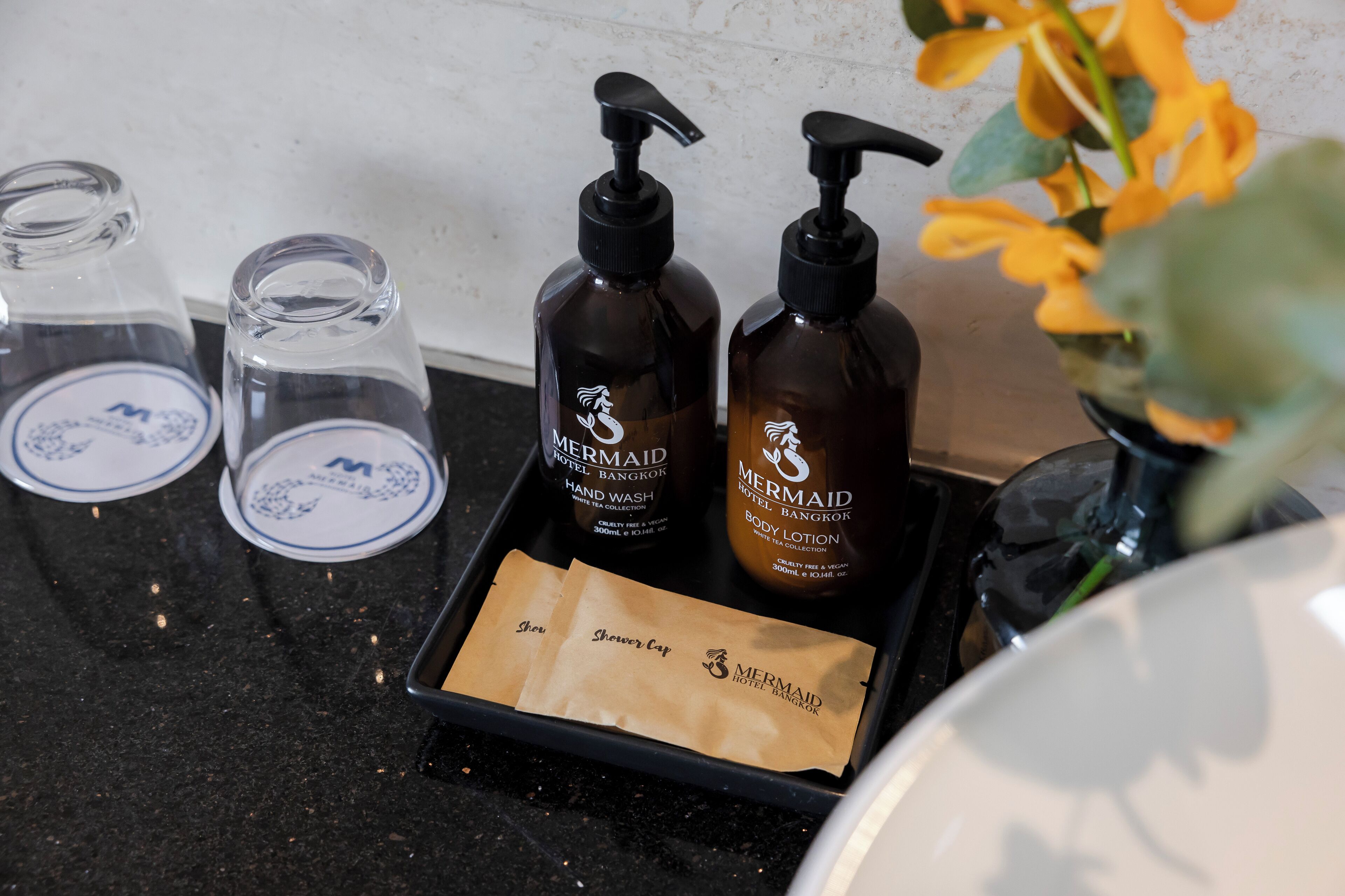 Ein Badezimmer-Waschtisch mit zwei Flaschen Mermaid Hotel Bangkok Körperlotion, einer Glasflasche und einem braunen Papiertuch.