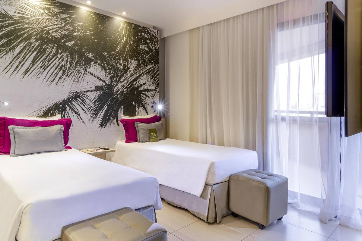 A hotel room with two beds, a large wall mural, a TV, and a window with sheer curtains.