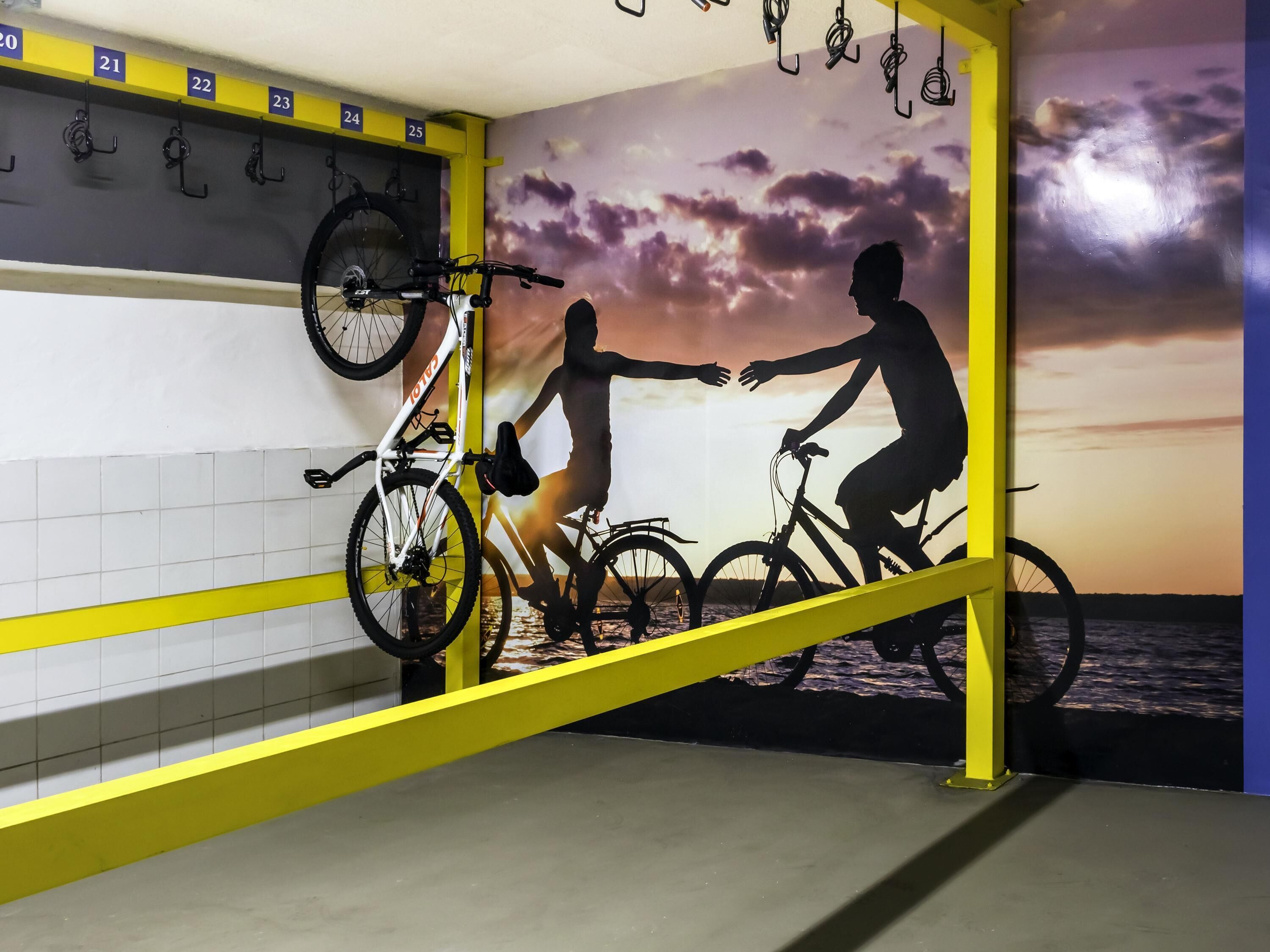A bike rack with numbered hooks, a wall mural of cyclists, and a yellow safety barrier.