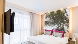 A modern hotel room with a large bed, a flat-screen TV mounted on the wall, a bedside table with a lamp, and a large window with sheer curtains. - Mercure Salvador Boulevard (Salvador)
