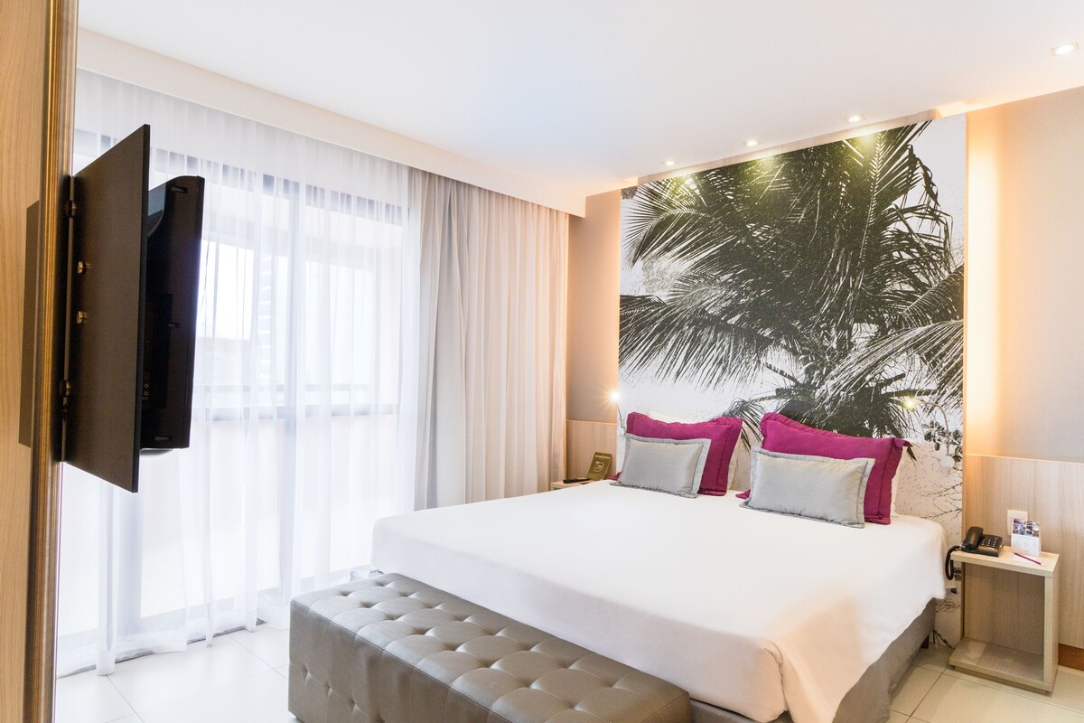 Una habitación de hotel moderna con una cama grande, un televisor de pantalla plana montado en la pared, una mesita de noche con lámpara y un ventanal con cortinas translúcidas.