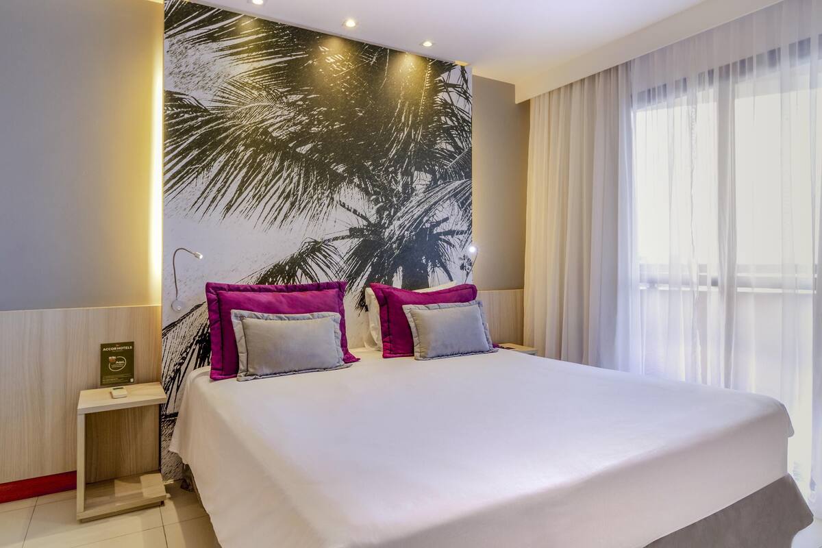 A hotel room with a large bed, two pillows, a nightstand, a wall mural, and a window with curtains.