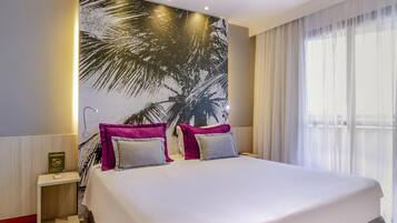 A hotel room with a large bed, two pillows, a nightstand, a wall mural, and a window with curtains.