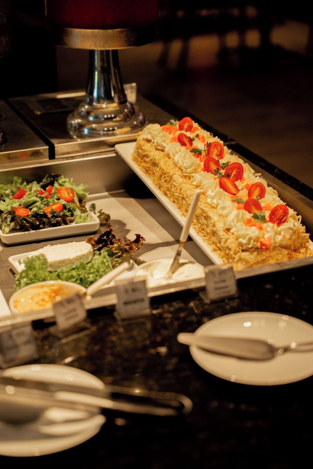 A buffet with a variety of dishes including a large rectangular cake with whipped cream and cherry tomatoes, a salad with lettuce and tomatoes, and other food items.