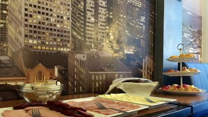 A breakfast buffet with ham, eggs, and fruit on a table near a window with a cityscape mural.