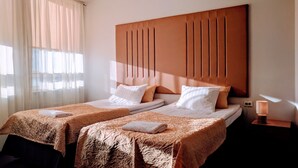 A hotel room with two beds, a wooden headboard, a bedside table with a lamp, and sheer curtains.