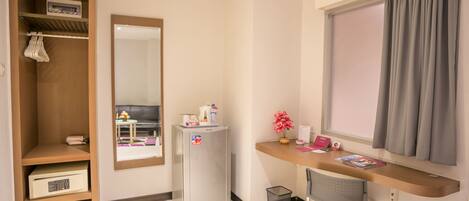 A compact hotel room with a wooden wardrobe, a small refrigerator, a desk with a chair, and a window with curtains.