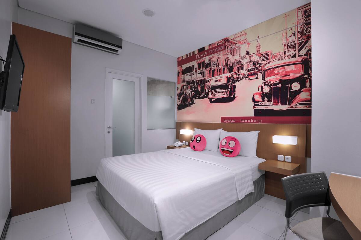 A hotel room with a bed, a nightstand, a chair, a wall-mounted TV, and a poster of a vintage car scene.