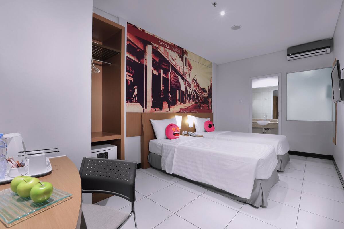 A hotel room with a bed, bedside tables, a desk with apples, a chair, a poster, and an air conditioning unit.