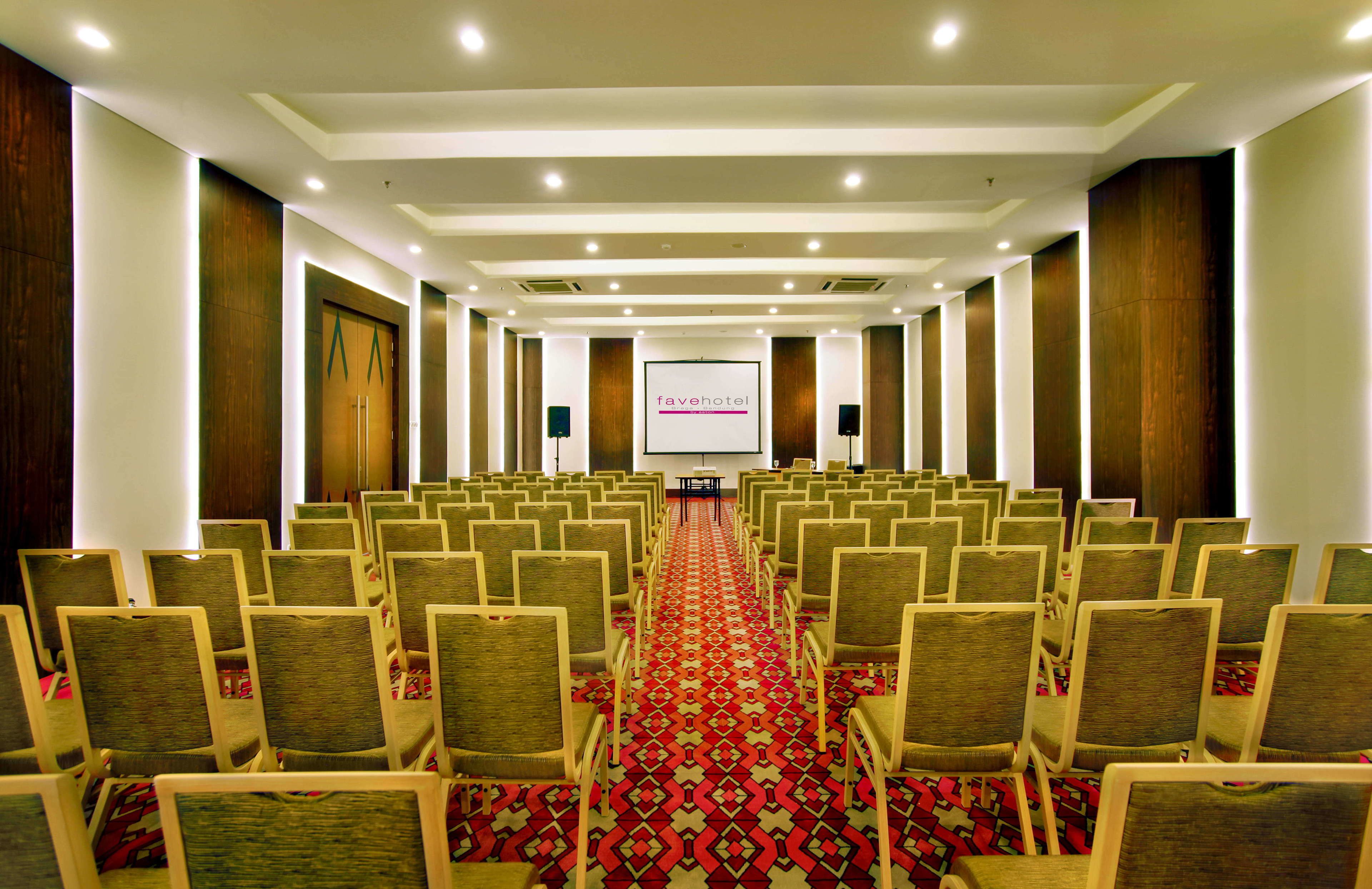 Une salle de conférence avec des rangées de chaises, une moquette rouge à motifs et un écran de projection affichant le nom « favehotel ».