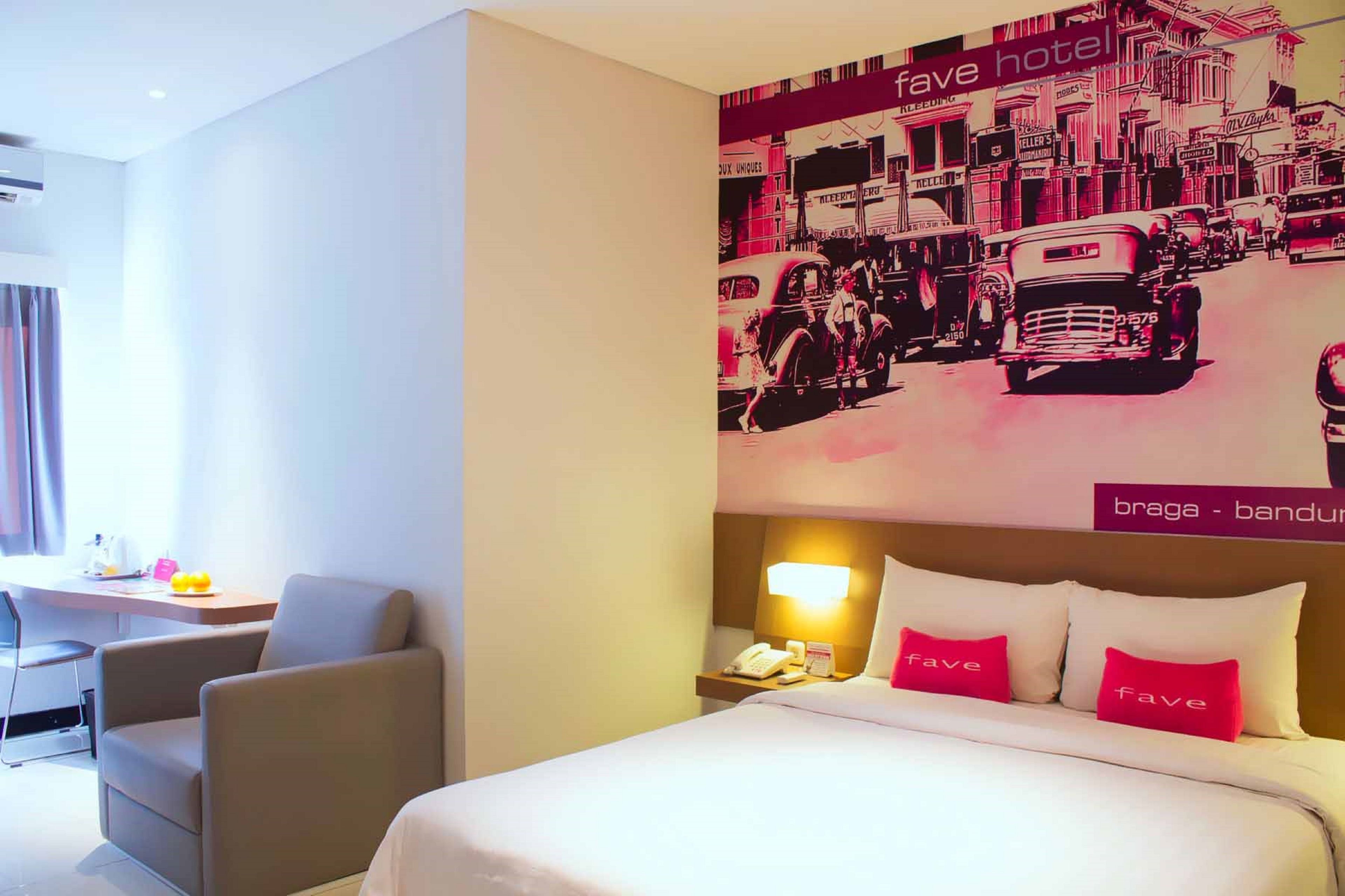 Habitación de hotel con cama, escritorio y silla. Se observa una pared con una ilustración de un coche antiguo y un edificio.