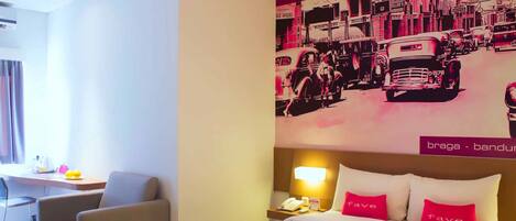 Habitación de hotel con cama, escritorio y silla. Se observa una pared con una ilustración de un coche antiguo y un edificio.