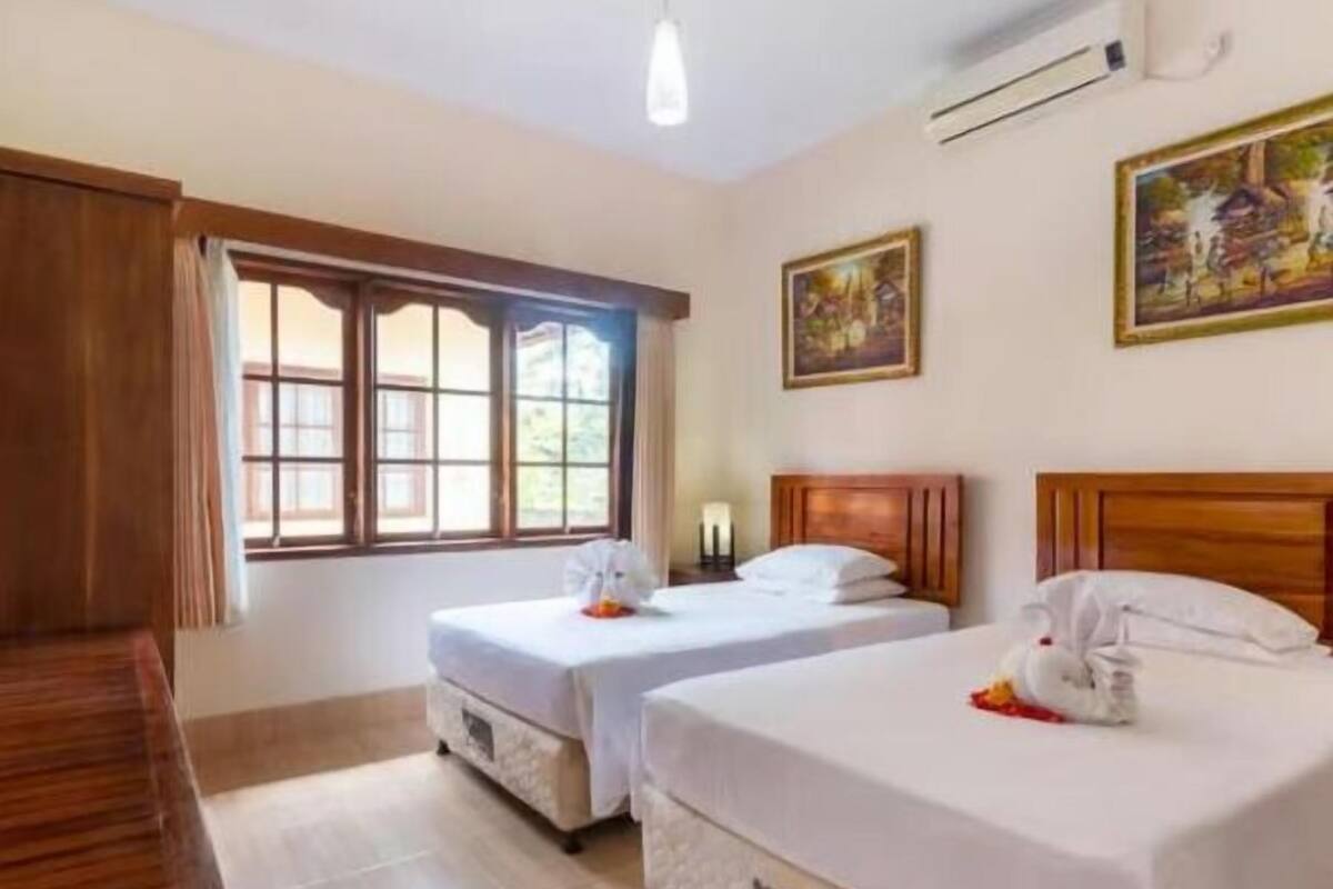 Een hotelkamer met twee bedden, een raam met gordijnen, een houten bankje en twee schilderijen aan de muur.