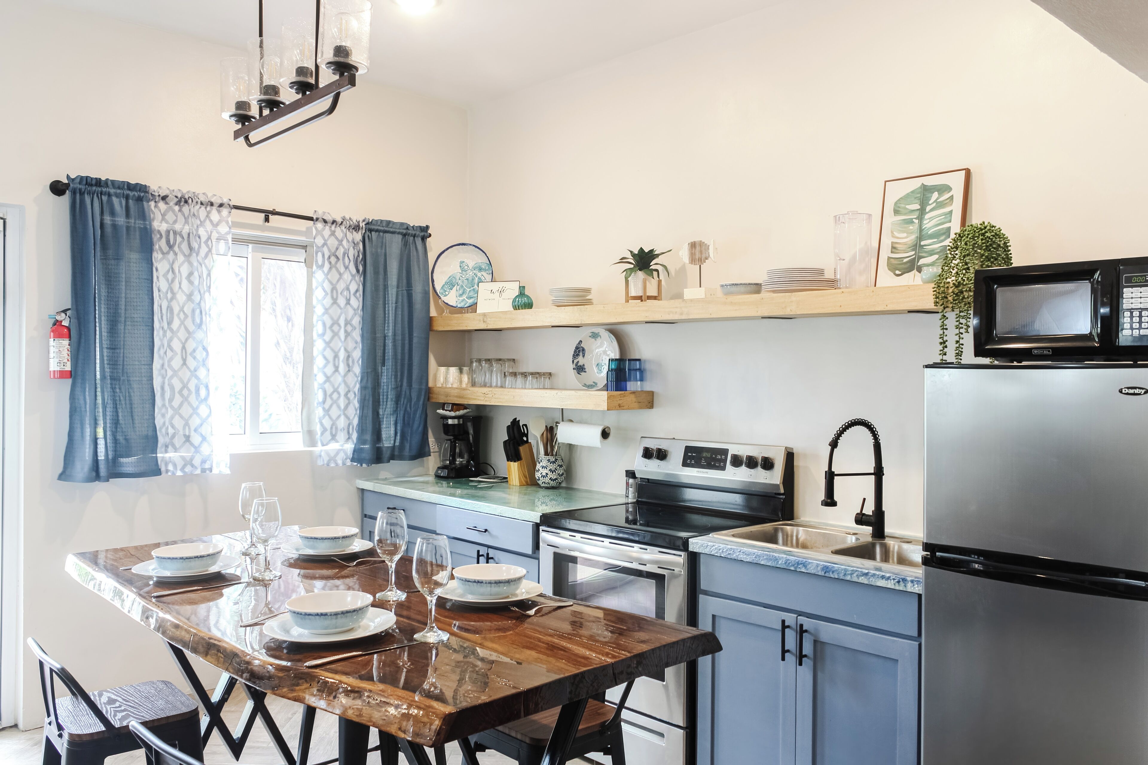 Una cocina moderna con una mesa de comedor de madera preparada para cuatro personas, electrodomésticos de acero inoxidable y una ventana con cortinas azules.