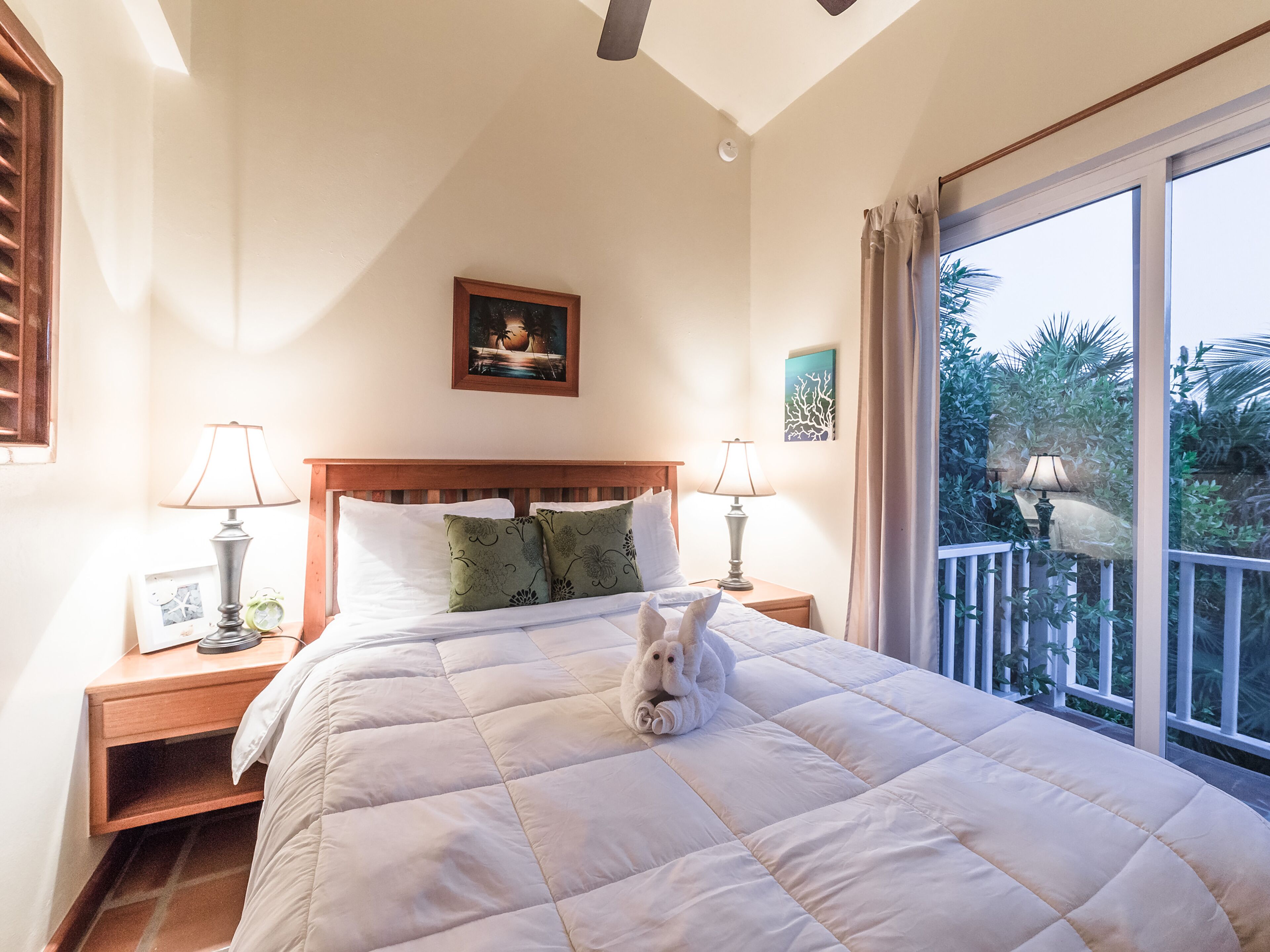 A bedroom with a large bed, two bedside tables with lamps, a framed picture on the wall, and a view of greenery through a balcony.