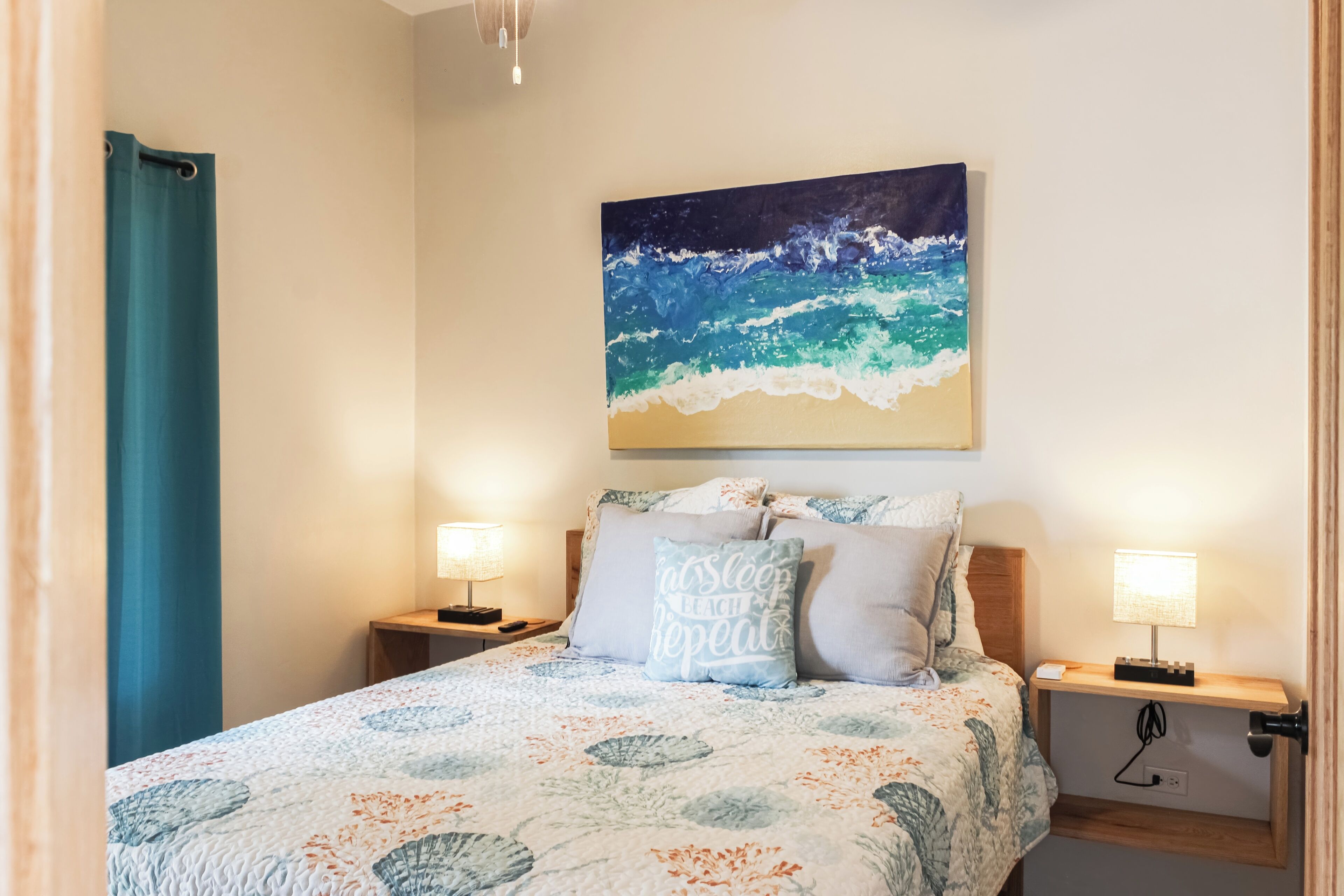 A bedroom with a bed, two bedside tables with lamps, a wall painting of ocean waves, and a blue curtain.