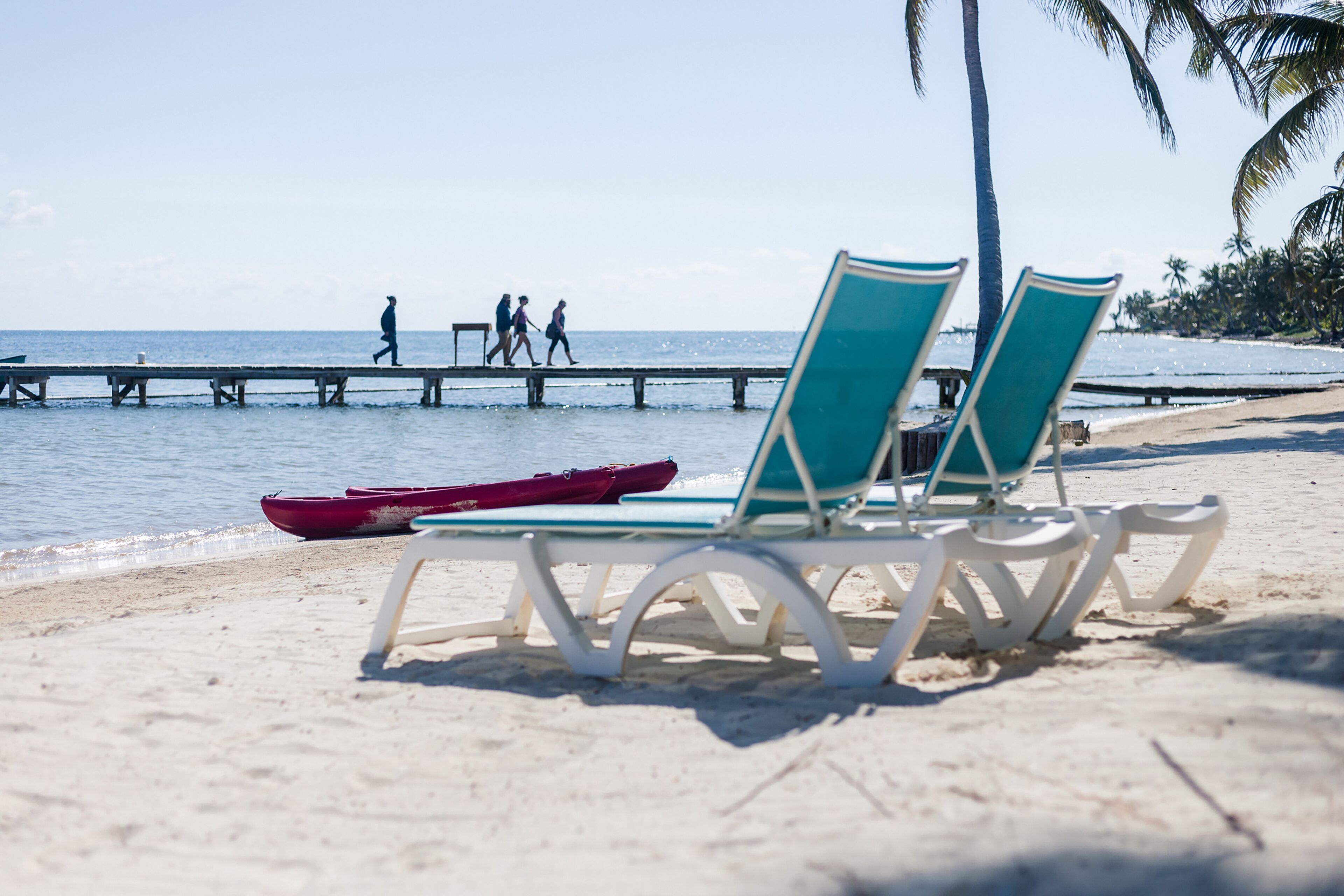 Ein Strand mit zwei weißen Liegestühlen, einem roten Boot und einer Mole, auf der Menschen spazieren.