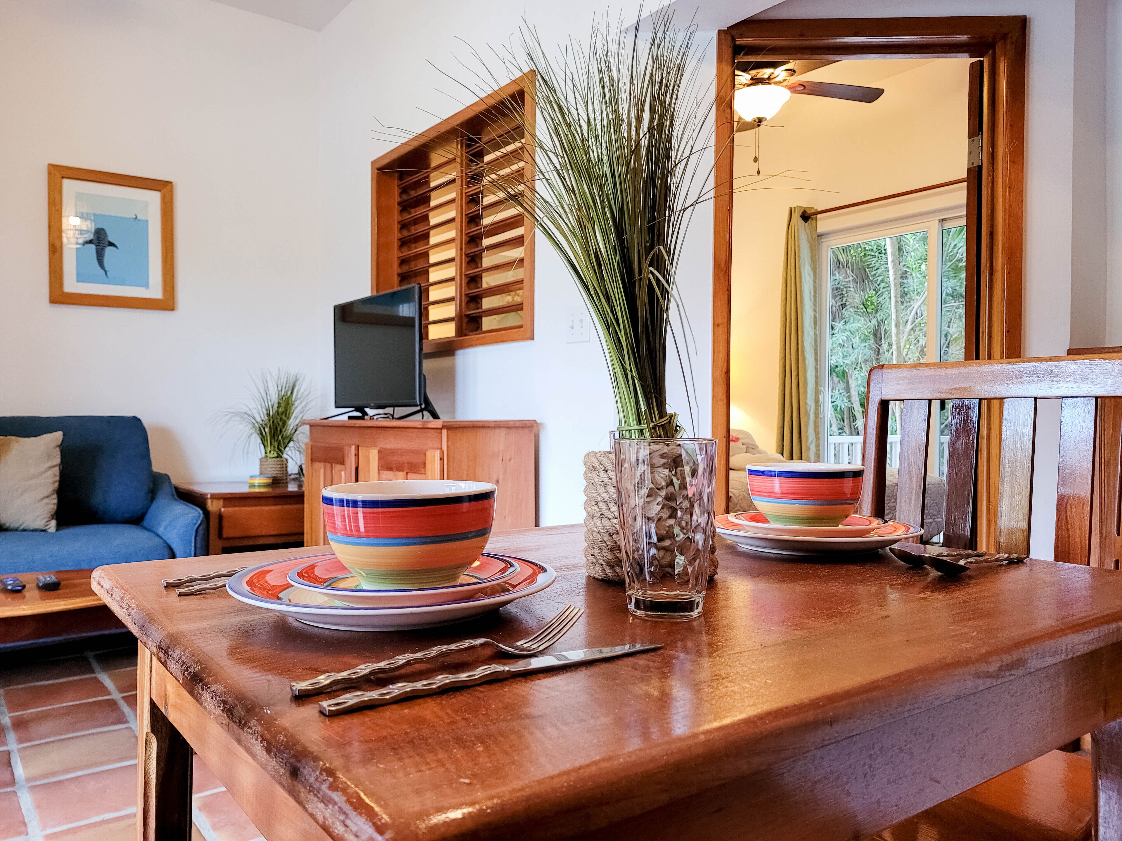 A dining table with a bowl, plates, and cutlery. There is a television on a wooden cabinet, a blue sofa, and a window with curtains.