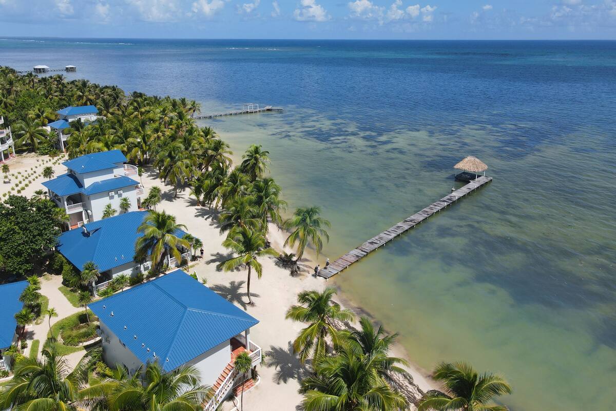Un resort con varios edificios de techos azules, una playa, palmeras y un muelle que se adentra en el océano.