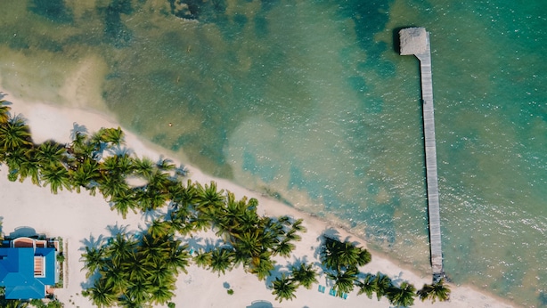 Eine Luftaufnahme eines Resorts mit mehreren Gebäuden mit blauen Dächern, einer langen Pier, die ins Wasser ragt, und einem Sandstrand, gesäumt von Palmen.