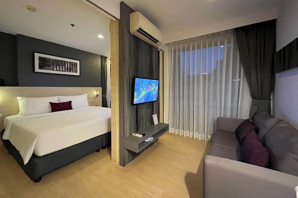 A hotel room with a bed, a TV, a sofa, and a large window with curtains.