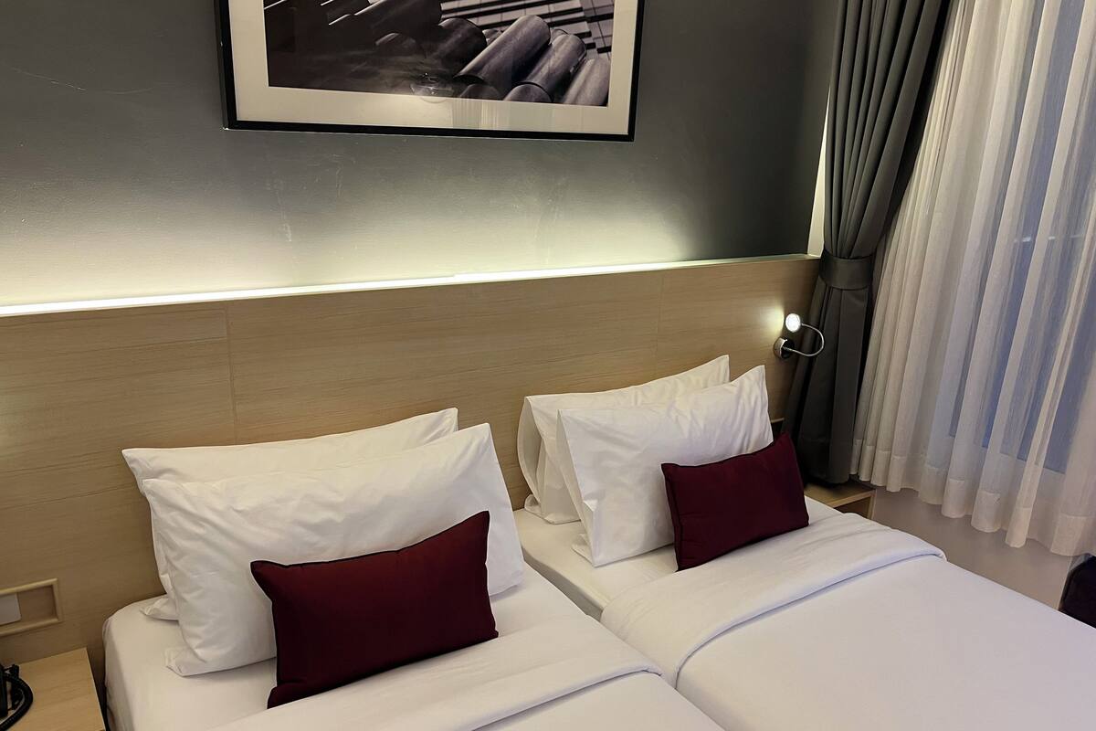 A hotel room with two beds, each with white bedding and a single dark red pillow. A framed picture hangs on the wall above the beds, and a nightstand with a telephone is visible to the left.