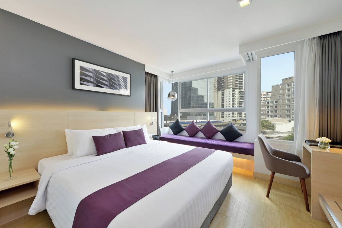 A modern hotel room with a large bed, a desk, a chair, a window with a city view, and a bedside table with a vase of flowers.